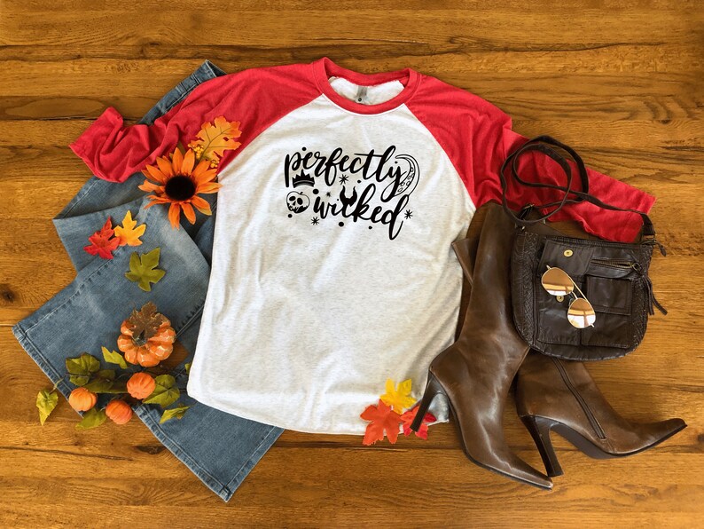 May include: A white and red baseball tee shirt with the text "perfectly wicked" in black. The shirt is styled with a pair of blue jeans, a brown leather bag, and brown leather boots.