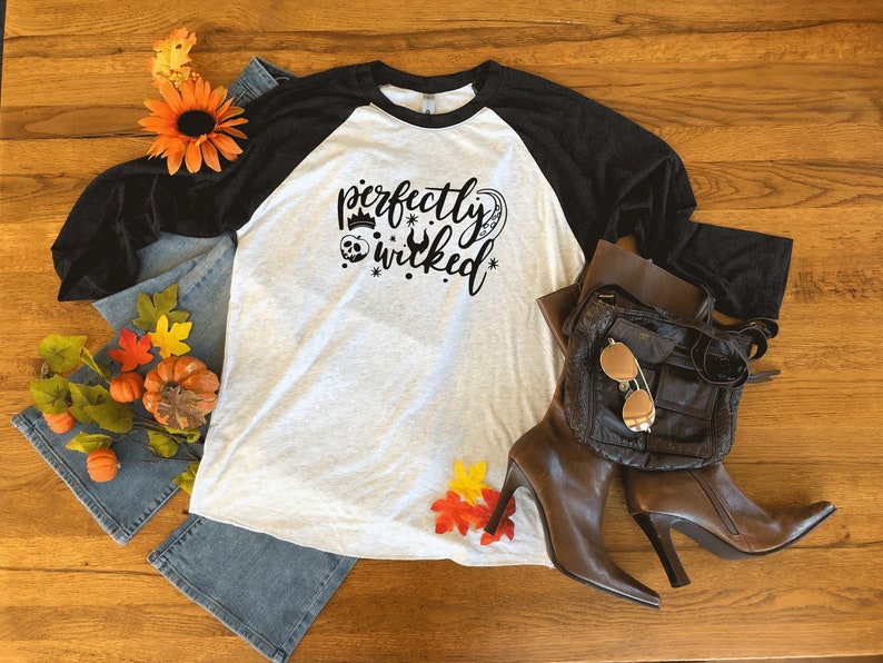 May include: A white and black baseball tee shirt with the text "perfectly wicked" in black script. The shirt has a black sleeve and a white sleeve. The text is accompanied by a crown, a skull, and a heart.