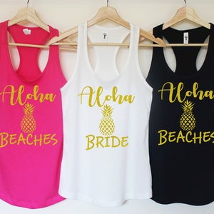Puede incluir: Tres camisetas sin mangas con el texto "Aloha" y un gráfico de piña. Las camisetas sin mangas son de color rosa, blanco y negro. El texto en la camiseta sin mangas rosa dice "Aloha Beaches", el texto en la camiseta sin mangas blanca dice "Aloha Bride" y el texto en la camiseta sin mangas negra dice "Aloha Beaches".