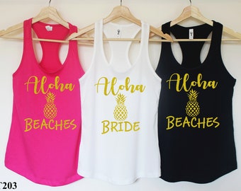 May include: Three tank tops with the text "Aloha" and a pineapple graphic. The tank tops are pink, white, and black. The text on the pink tank top says "Aloha Beaches", the text on the white tank top says "Aloha Bride", and the text on the black tank top says "Aloha Beaches".