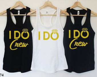 May include: Three tank tops with the text "I DO Crew" in gold glitter lettering. The middle tank top is white, and the two outer tank tops are black. Each tank top features a gold glitter diamond ring.