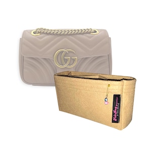 Peut inclure: Un organisateur de sac en feutre beige avec plusieurs compartiments intérieurs, une petite épingle à nourrice dorée avec une breloque œil porte-bonheur rose, et une étiquette en tissu noir indiquant Felt Bag Organizer by Seda Bektas.