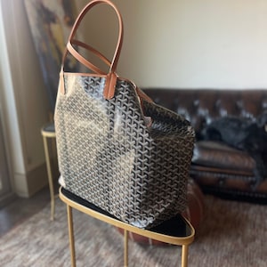 Puede incluir: Un bolso tote marr&oacute;n y negro con asas de cuero marr&oacute;n. El bolso est&aacute; sobre un soporte de metal dorado con la parte superior negra.