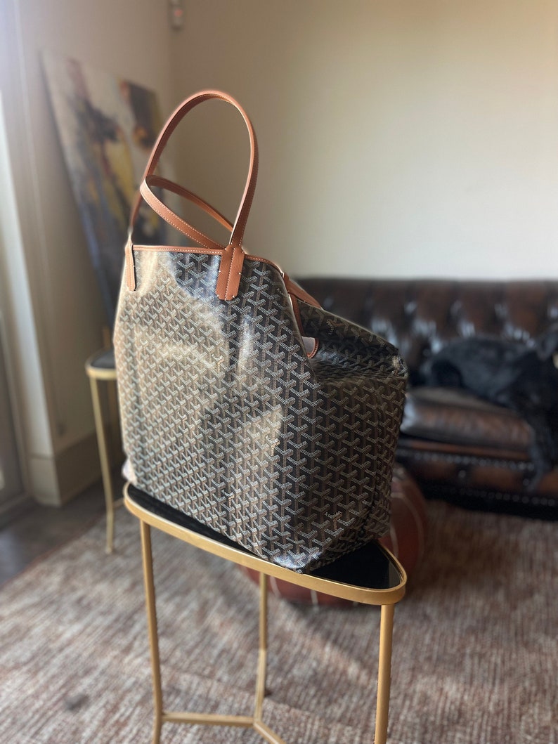 Puede incluir: Un bolso tote marr&oacute;n y negro con asas de cuero marr&oacute;n. El bolso est&aacute; sobre un soporte de metal dorado con la parte superior negra.