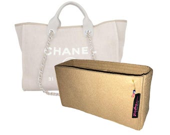 May include: A beige felt bag organizer with a black side tag reading Felt Bag Organizer and a small evil eye charm, positioned in front of a cream-colored canvas tote bag featuring the text CHANEL and a silver chain strap detail.