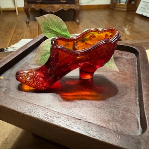 Puede incluir: Un zapato de cristal rojo rubí vintage con intrincados diseños florales en relieve. El zapato decorativo está sobre una bandeja de madera, mostrando su tacón alto y su parte superior abierta. El cristal tiene un cálido brillo ámbar.