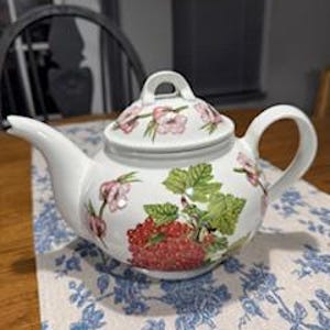 Puede incluir: Tetera de cerámica blanca con tapa, con un diseño floral con bayas rojas y hojas verdes. La tetera tiene un pico curvo y un asa. Se encuentra sobre un mantel con estampado floral azul y blanco. Un artículo de cocina clásico.