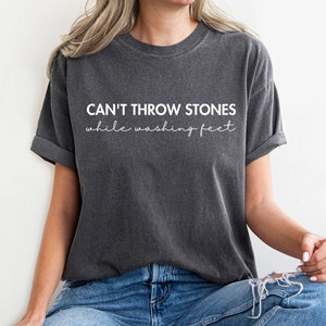 May include: Dark gray t-shirt with the text "CAN'T THROW STONES while washing feet" in white. The shirt has short sleeves and a relaxed fit. The model is wearing blue jeans.
