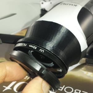 Op de afbeelding: Een zwart-witte Parrot-drone met een 52 mm UV-filter bevestigd aan de cameralens. De drone heeft zwarte propellers en ligt op een tijdschrift. Het filter wordt opengehouden.