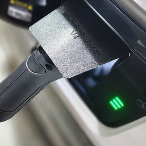 May include: A black electric vehicle charging cable plugged into a charging port with a green light indicating charging in progress. The charging port has a black cover with the text "WARNING HOT SURFACE" and the number "V4" printed on it.