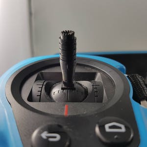 Peut inclure: Gros plan sur un joystick noir avec une base bleue. Le joystick est centré sur l'image et possède un bouton noir avec une surface texturée. La base du joystick est de couleur bleu vif.