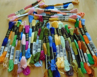 May include: A collection of colorful embroidery floss skeins in various shades of blue, green, yellow, pink, and orange. Each skein is labeled with a number and wrapped in a black and white label. The floss is arranged on a light-colored wooden surface.