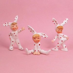 May include: Three porcelain figurines of children dressed in white bunny costumes with red polka dots. The figurines are posed in different positions, with one standing and two sitting.