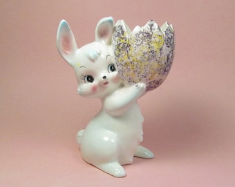 May include: A white ceramic bunny figurine holding a speckled egg-shaped planter. The bunny has blue eyes and pink ears. The planter is decorated with purple, yellow, and white speckles.