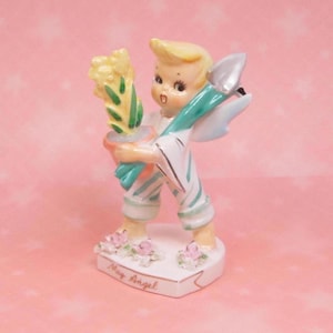 May include: A vintage ceramic figurine of a cherubic angel holding a bouquet of yellow flowers and a silver trowel. The angel wears a white outfit with blue and white striped accents. The base is white with pink flowers and the words "May Angel".