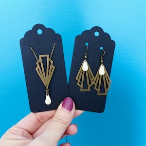 Può includere: Un set di gioielli dorati composto da una collana e orecchini. La collana ha un ciondolo a forma di ventaglio art déco con una pietra bianca nella parte inferiore. Anche gli orecchini hanno la forma di un ventaglio art déco con pietre bianche.