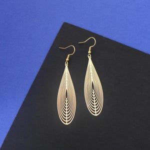 Puede incluir: Un par de pendientes colgantes de color dorado con un diseño geométrico en forma de hoja. Los pendientes están hechos de metal fino y tienen un aspecto delicado y aireado.