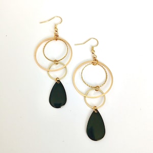 Peut inclure: Une paire de boucles d'oreilles pendantes dorées au design multicouche. Chaque boucle se compose de deux cercles dorés entrelacés, l'un lisse et l'autre texturé, suspendus à un crochet et terminés par une breloque en forme de goutte noire.