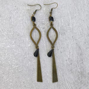 Puede incluir: Un par de pendientes colgantes con estética vintage. Cada pendiente presenta una cuenta cuadrada negra, un elemento ovalado color latón, una cuenta negra en forma de lágrima y un colgante largo y estrecho color latón. Los pendientes cuelgan de un gancho simple.