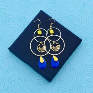 Puede incluir: Pendientes de aro dorados con colgantes en forma de lágrima azul y amarillo. Los pendientes tienen un diseño geométrico con un círculo y un círculo más pequeño en el interior.