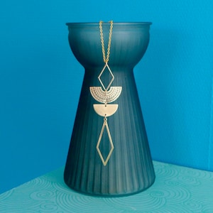 Può includere: Una collana d'oro con un design geometrico pende da un vaso blu scuro. La collana presenta un grande cerchio aperto con una piccola forma di diamante sottostante. La collana è su una superficie con un motivo blu e bianco.