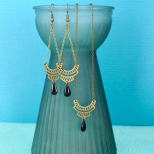Op de afbeelding: Een set gouden oorbellen en een ketting met zwarte traanvormige stenen. De oorbellen en ketting hebben een geometrisch ontwerp met een waaiervormig patroon.