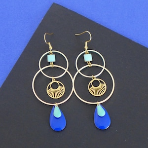 Può includere: Orecchini a cerchio dorati con un ciondolo a goccia blu e turchese, un ciondolo quadrato più piccolo e un design decorativo a forma di sole.