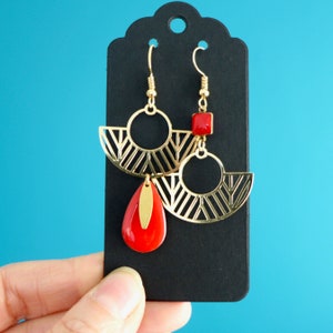 Puede incluir: Un par de pendientes de oro con diseños geométricos y detalles rojos. Los pendientes tienen un diseño en forma de abanico con un pequeño cuadrado rojo y una piedra roja en forma de lágrima.