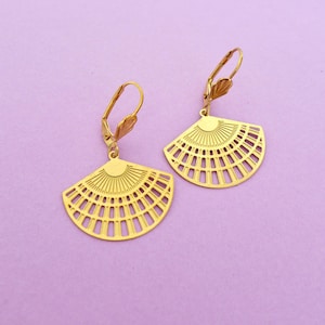 Może przedstawiać: Para złotych kolczyków w kształcie wachlarza z zapięciem zatrzaskowym. Kolczyki mają szczegółowy, ażurowy wzór z motywem słońca u góry. Tło jest jednolite, jasnofioletowe.