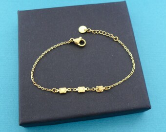 Puede incluir: Una delicada pulsera dorada con tres dijes rectangulares decorativos. La pulsera tiene una cadena con una longitud ajustable y un cierre de mosquetón. La joya se exhibe en una caja negra sobre un fondo azul.