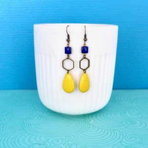 May include: A pair of earrings with a geometric design. Each earring has a dark blue square, a bronze-coloured hexagon, and a yellow teardrop shape. The earrings hang from bronze-coloured ear wires and are displayed in a white cup against a blue background.