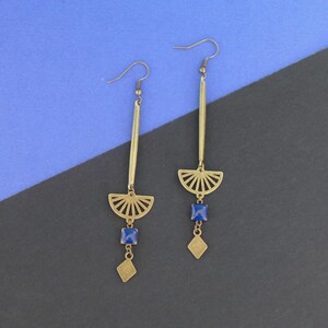 Peut inclure: Une paire de boucles d'oreilles dorées avec un design géométrique. Chaque boucle d'oreille présente un pendentif en forme d'éventail avec une pierre précieuse bleue et un plus petit pendentif en forme de diamant.