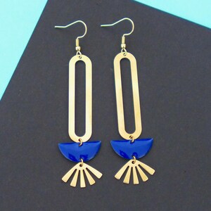 Peut inclure: Une paire de boucles d'oreilles géométriques dorées. Chaque boucle d'oreille présente une longue forme ovale en haut, un demi-cercle bleu au milieu et un motif en forme d'éventail en bas. Les boucles d'oreilles sont suspendues à des crochets dorés.