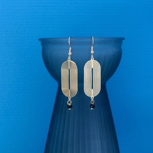 Peut inclure: Boucles d'oreilles ovales dorées avec un motif géométrique et des accents de perles noires.