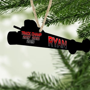 May include: Black silhouette of a race car with red text that reads "TRACK CHAMP 2017 2018 2019" and "RYAN" on a white background. The ornament is hanging from a brown string.