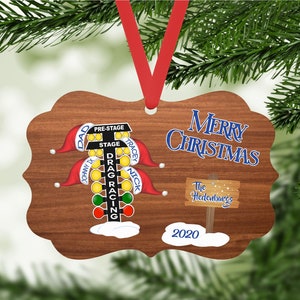 May include: A wooden Christmas ornament featuring a drag racing Christmas tree with red, green, and yellow lights. The ornament has the names "Dad", "Johnny Ty", "Tracey", and "Nick" on it. The ornament also has the text "Merry Christmas" and "The Hedenburgs" on it. The year "2020" is also on the ornament.