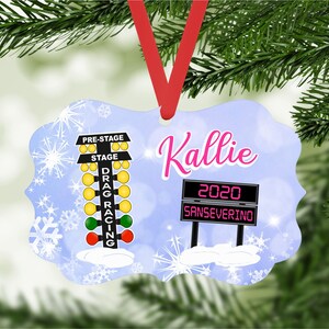 May include: A blue and white Christmas ornament featuring a drag racing Christmas tree with the words "Pre-Stage" and "Stage" and a sign that says "2020 Sanseverino". The ornament also has the name "Kallie" written in pink.