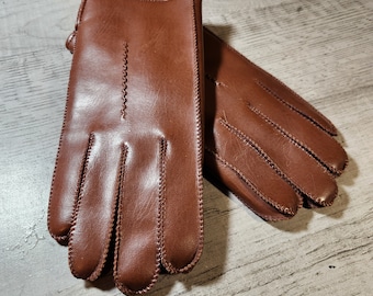May include: A pair of brown leather gloves with a stitched detail around the edges. The gloves are laid out flat on a wooden surface.