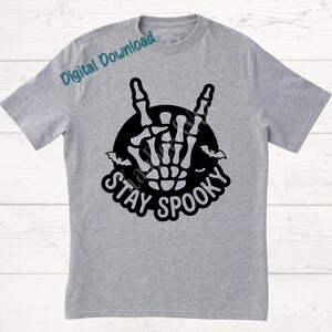 May include: A light gray t-shirt featuring a black graphic of a skeleton hand making a rock and roll gesture, surrounded by a black circle and bats. The words "STAY SPOOKY" are written below the graphic in a stylized font.