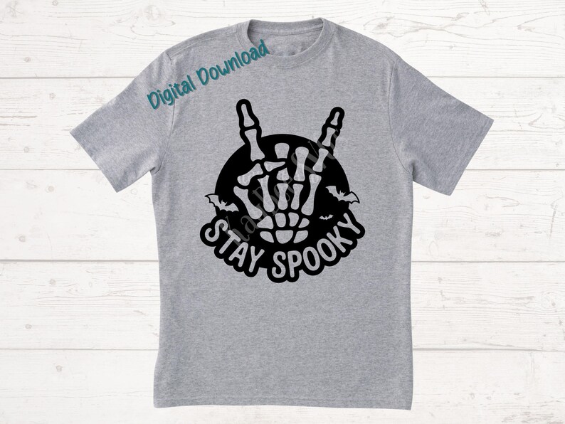 May include: A light gray t-shirt featuring a black graphic of a skeleton hand making a rock and roll gesture, surrounded by a black circle and bats. The words "STAY SPOOKY" are written below the graphic in a stylized font.