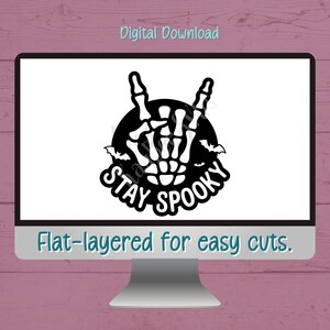 May include: A computer screen displays a black and white Halloween-themed graphic. The design features a skeleton hand making a rock-and-roll gesture, surrounded by bats and the words "STAY SPOOKY." The text "Digital Download" and "Flat-layered for easy cuts" are also visible.