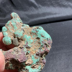 May include: A natural mineral specimen with a rough, irregular shape. The stone features a mix of colors, including shades of green, brown, and purple. The surface has a textured appearance, with areas of crystalline formations.