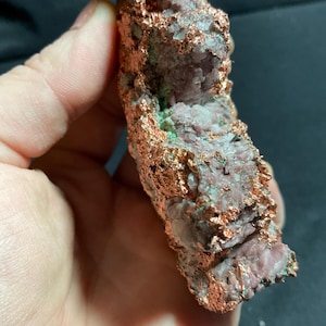 May include: A raw copper specimen with a rough, textured surface. The copper has a reddish-brown color with areas of green and pinkish-grey hues. The mineral appears to be a natural formation, possibly a geode or a similar type of rock.