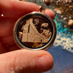 May include: A round, gold-colored locket pendant with a clear glass front. Inside, a wooden cutout of a dam is surrounded by small, gold-colored stones. The pendant is held by a hand.