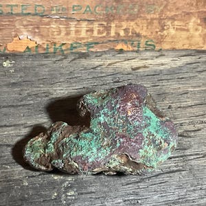 May include: A natural, irregularly shaped stone with a textured surface. The stone displays a mix of colors, including shades of green, brown, and reddish-brown. The stone is set against a weathered wooden background.