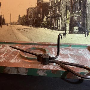 May include: An antique metal tool with a hook and handle, resting on a weathered, rectangular tray with green trim. The background features a sepia-toned vintage photograph of a street scene.