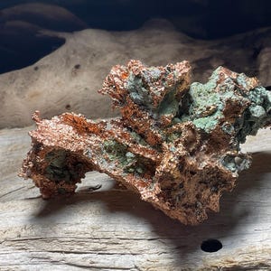 May include: A raw mineral specimen featuring a metallic copper-colored crystalline structure interspersed with patches of pale green oxidation. The formation has a jagged, irregular, and porous texture with sharp edges and deep crevices throughout the piece.