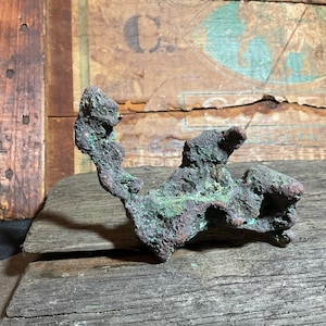 May include: A weathered, irregularly shaped object with a textured surface. The object displays a mix of colors, including green, brown, and reddish hues. It rests on a rough wooden surface, with a backdrop of aged wood.