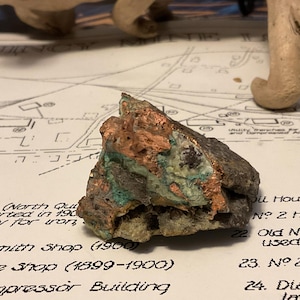 May include: A close-up of a rough, dark gray rock with patches of copper and green. The rock has a natural, unpolished look.