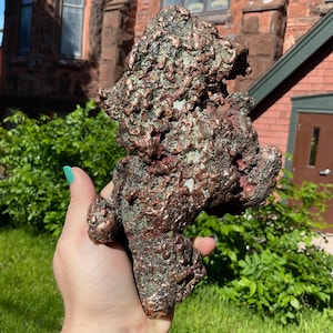 May include: A raw, naturally formed copper specimen featuring a highly textured, metallic surface with a reddish-brown luster and patches of green oxidation. The mineral chunk has an irregular, jagged shape with deep crevices and protruding nodules.